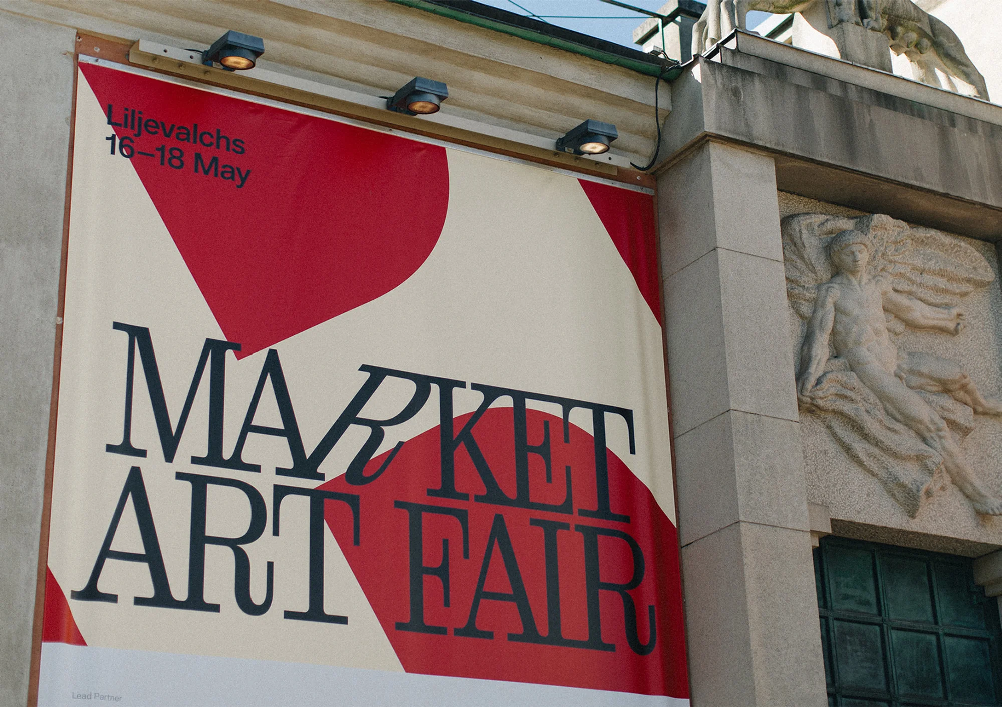 Market Art Fair banner hanging on stone building featuring modern typography and red and white geometric shapes and an angel sculpture.