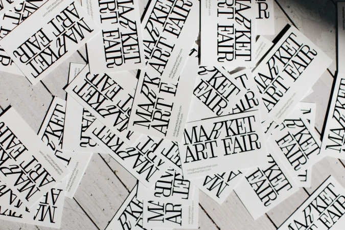 Multiple white cards scattered on a grey wooden surface. The cards are printed with the text 'Market Art Fair' in bold, black lettering.