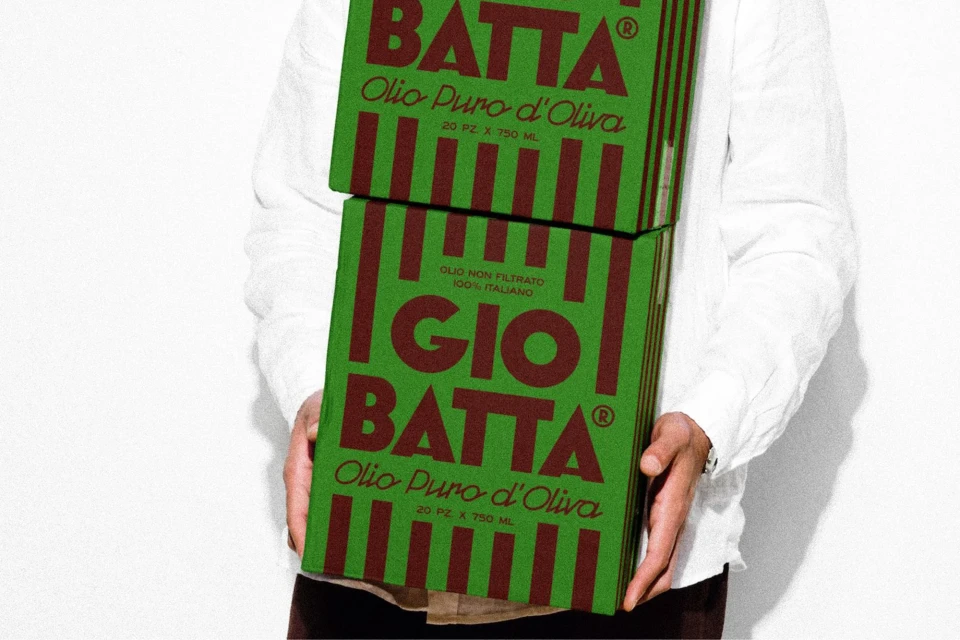 Person holds two Gio Batta olive oil boxes in a studio setting. The packaging is green with brown stripes and Italian text.