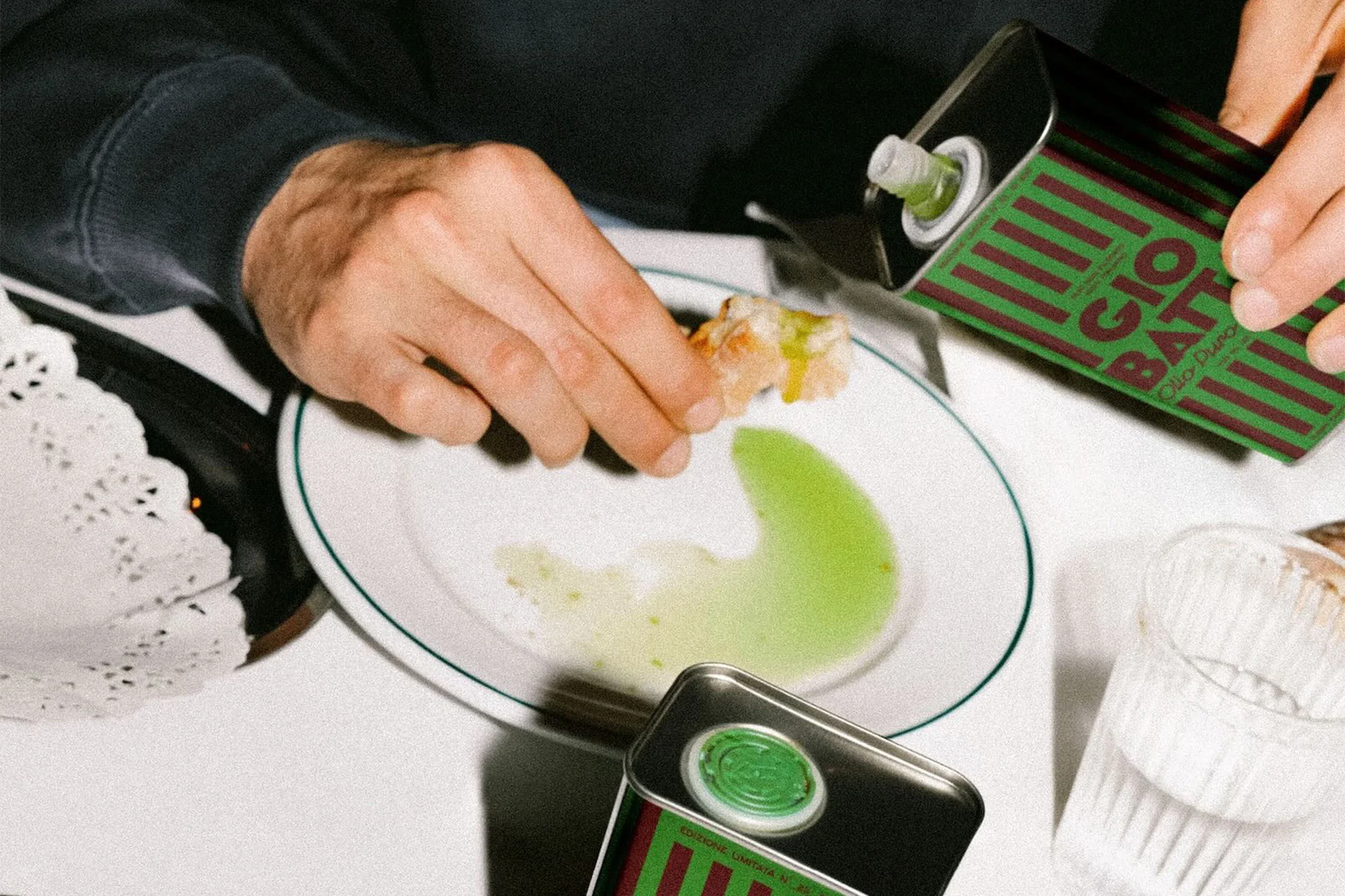 Person dips bread in olive oil from Giobatta tin can on a white plate. A glass and lace doily are on the white tablecloth.