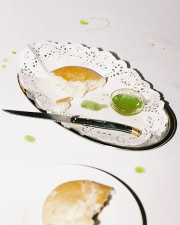 Overhead shot of bread and green sauce on a silver tray with a doily, sitting on a white tablecloth. A knife and another plate are visible.