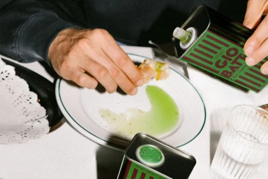 Close-up of hands dipping bread into bright green olive oil poured from a Giobatta tin onto a white plate.