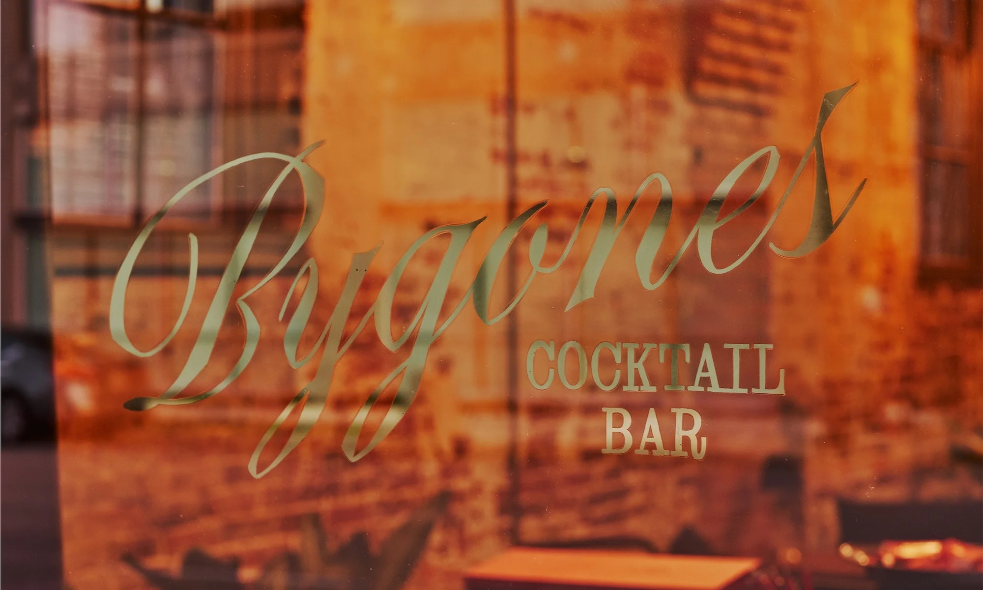 Close-up shot of the vintage gold lettering 'Bygone's COCKTAIL BAR' on a window, reflecting an orange brick building.