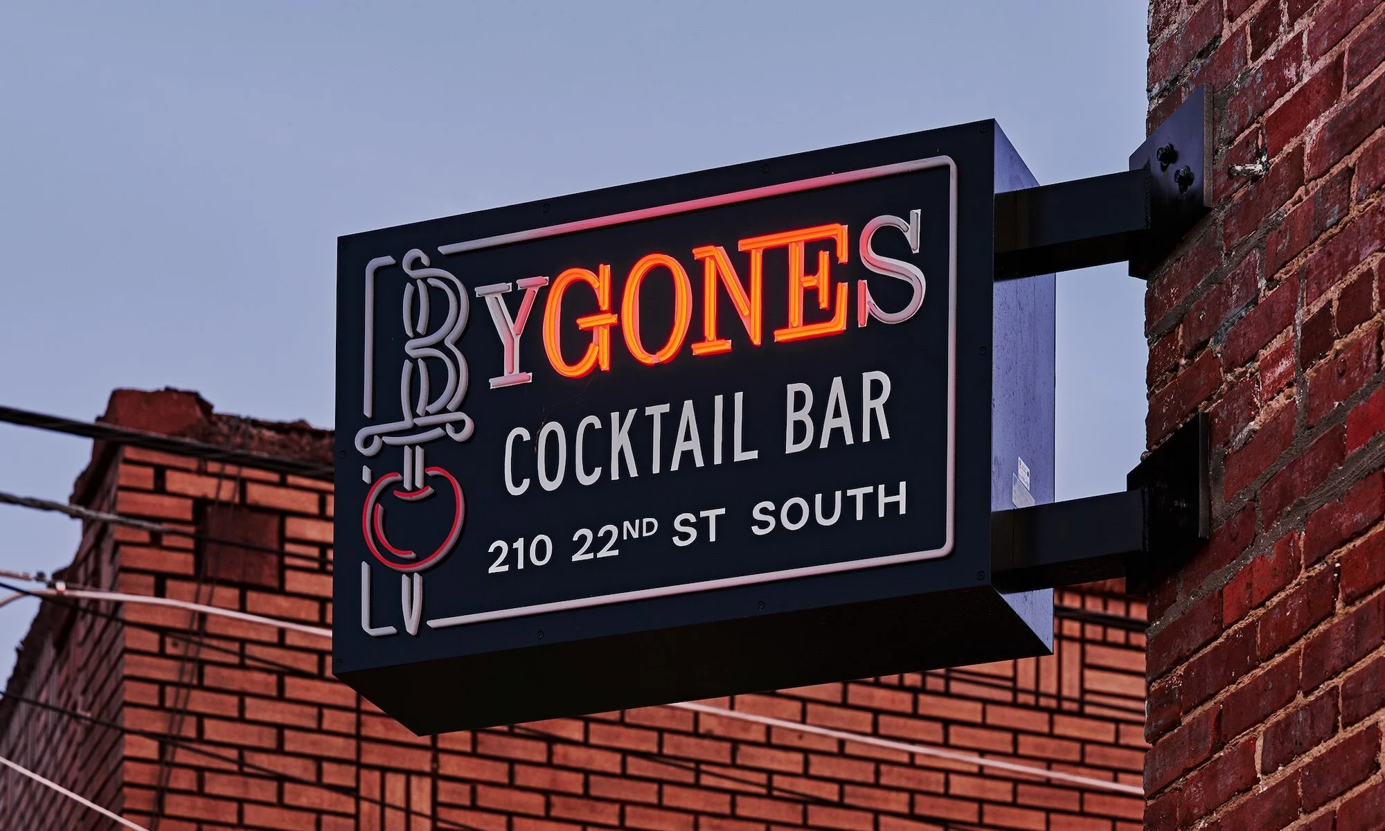 Illuminated sign for Bygones Cocktail Bar featuring orange and white neon lettering and a stylized cherry design, mounted on a brick building.