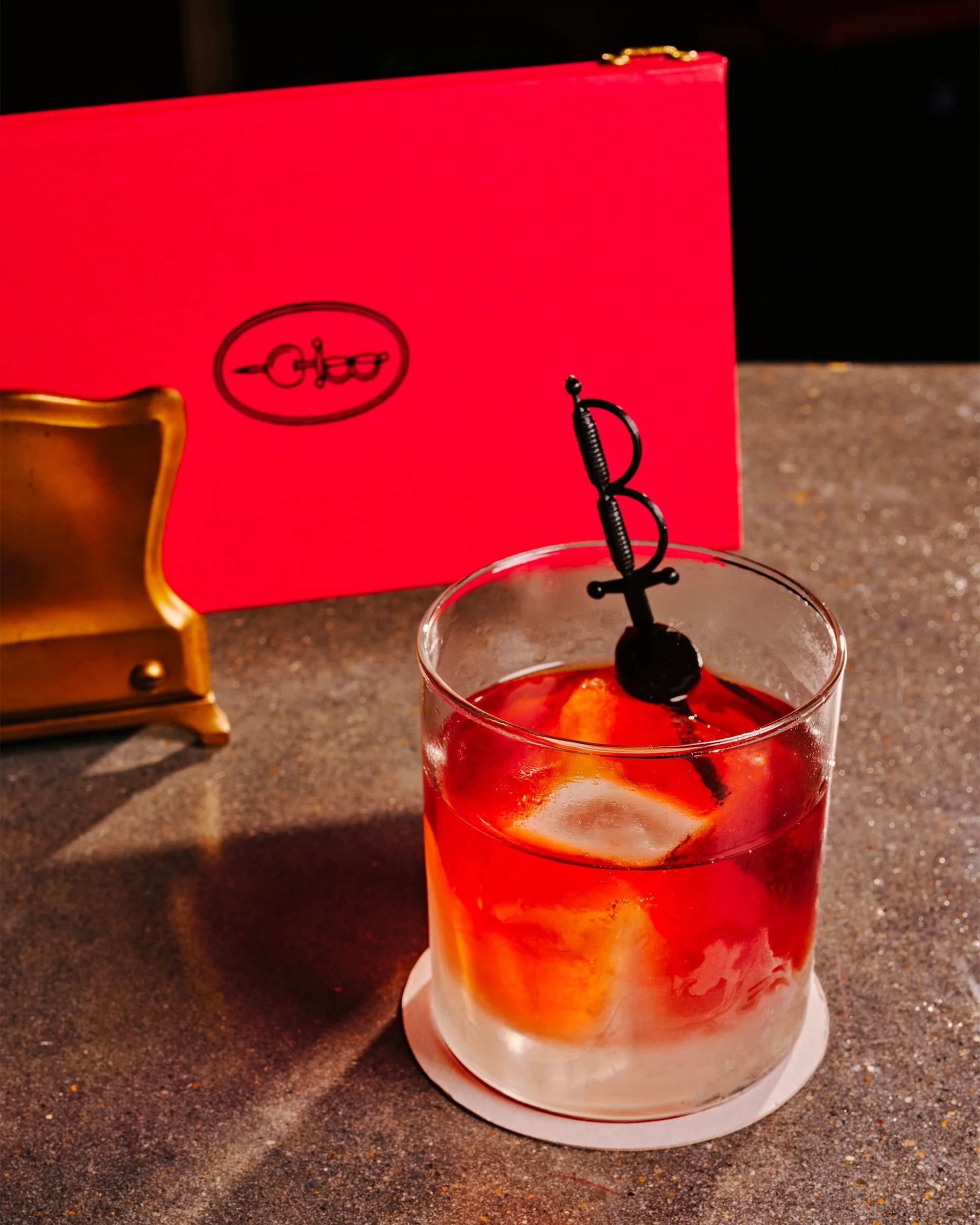 Elegant cocktail on a coaster next to gold object and red box featuring logo on a textured grey table inside a bar setting.