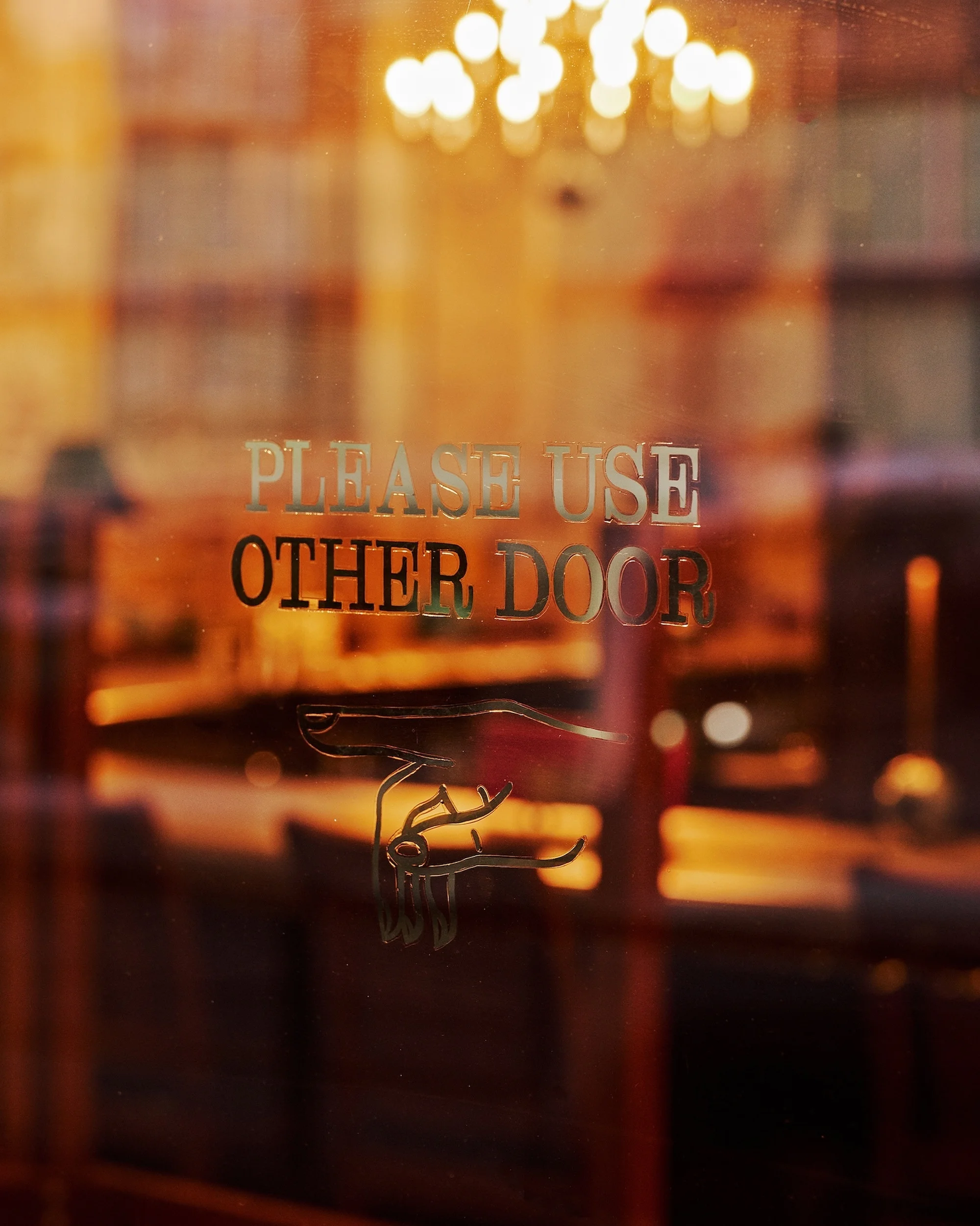 A glass door with an etched design of a hand pointing towards text that reads 'Please Use Other Door' in a warm, blurred interior.