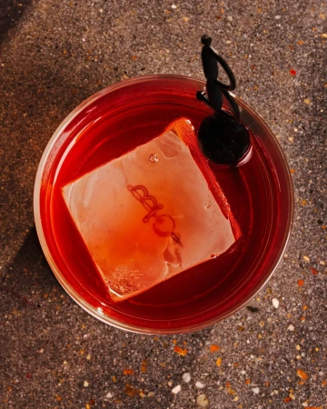 Overhead view of a red cocktail in a clear glass with a large ice cube, garnished with a black cocktail pick and orange peel on stone.