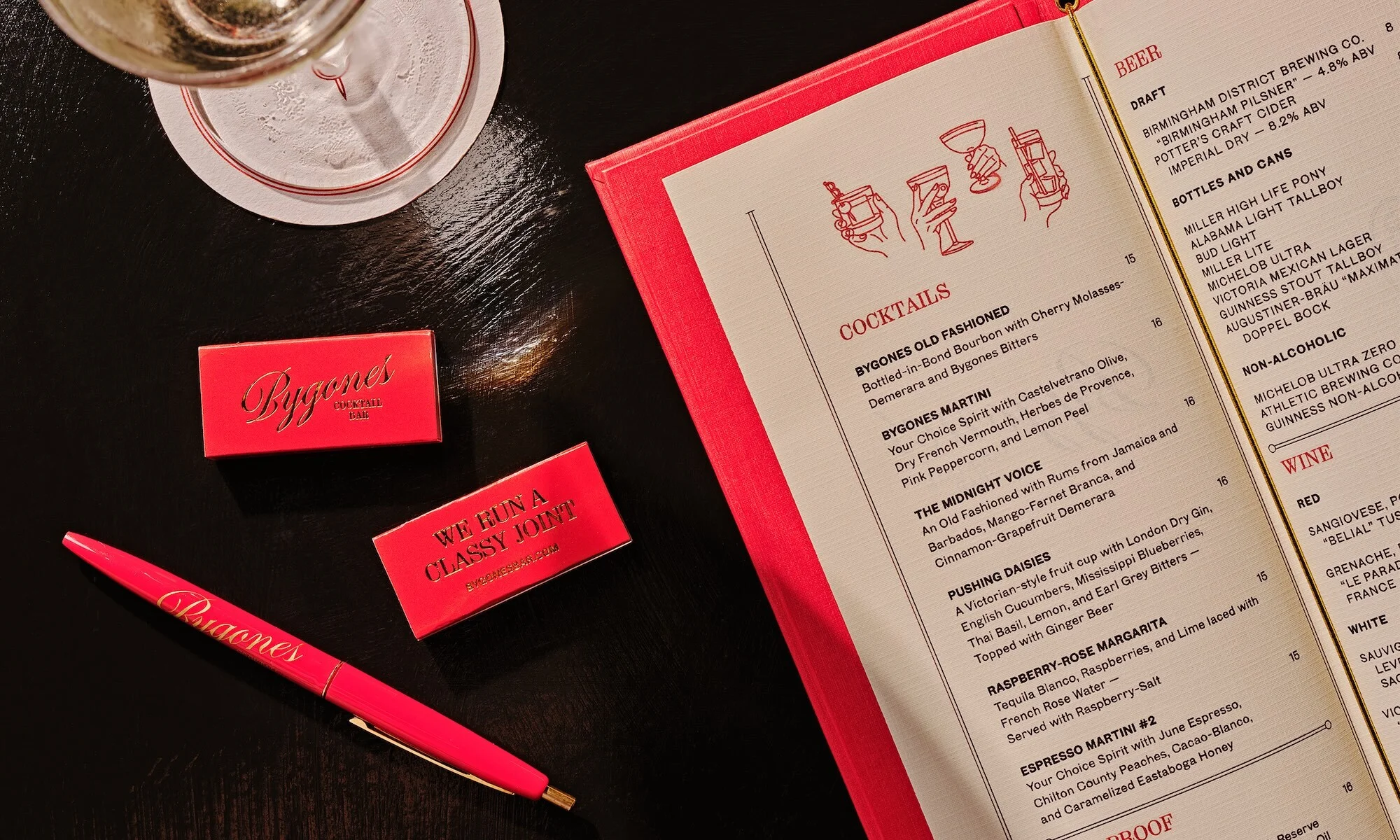 Overhead shot of Bygones brand pen, matches, and menu with cocktail list on dark table with drink; pink, red, black, and white colors.