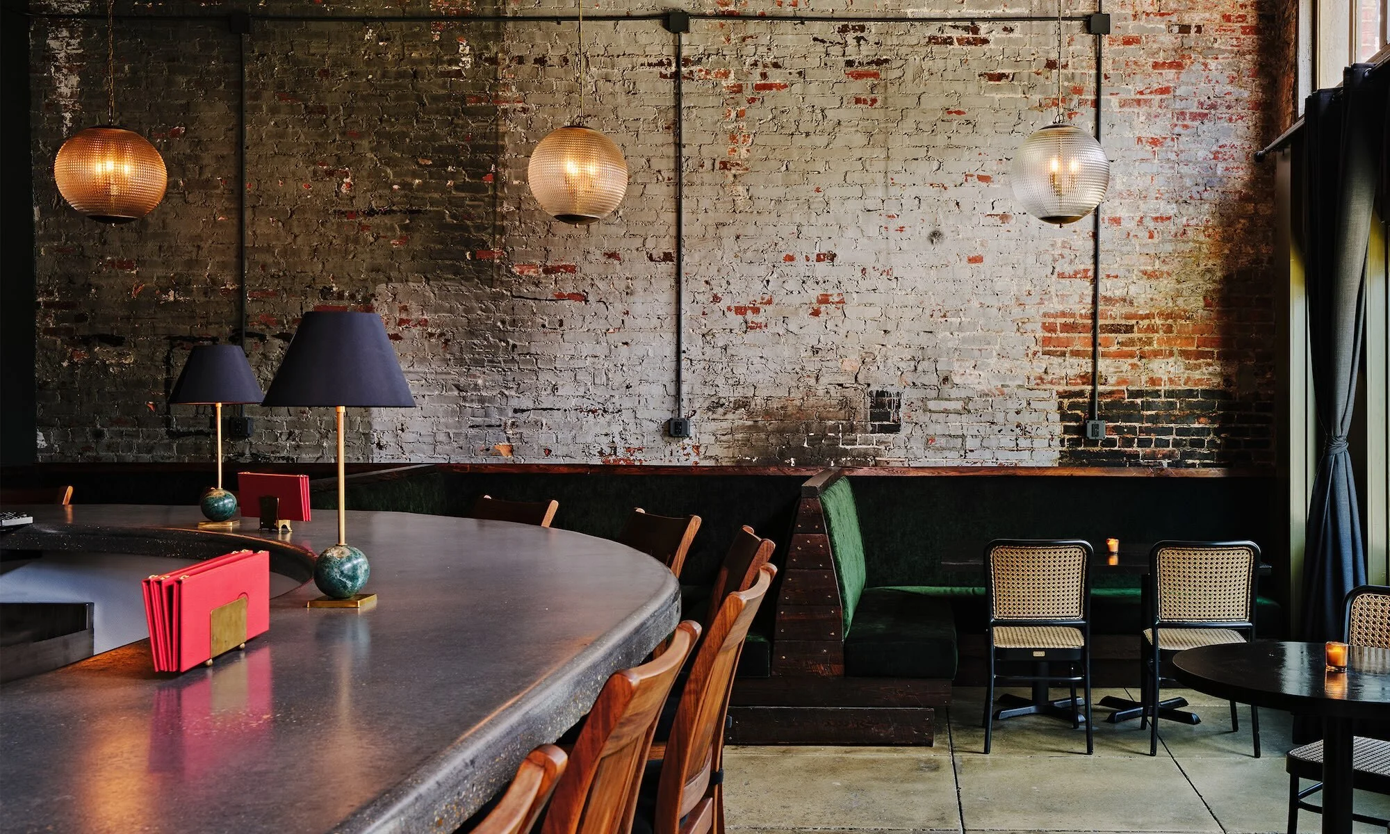 Interior view of a dimly lit bar with a weathered brick wall, globe pendant lights, and tables with chairs, creating a warm ambiance.