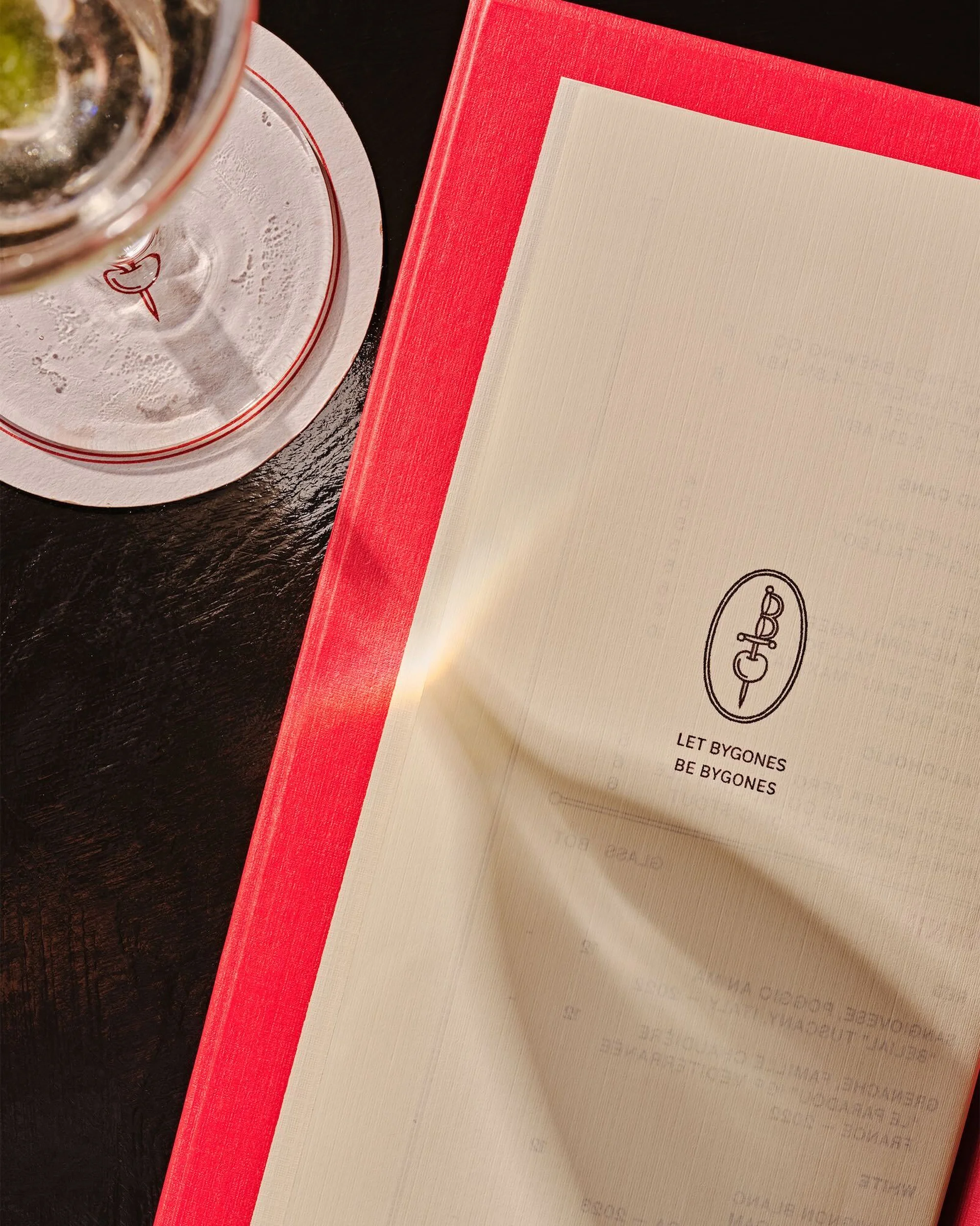 Overhead shot of a beverage glass with a coaster next to a red menu on a dark table, featuring minimal design and text.