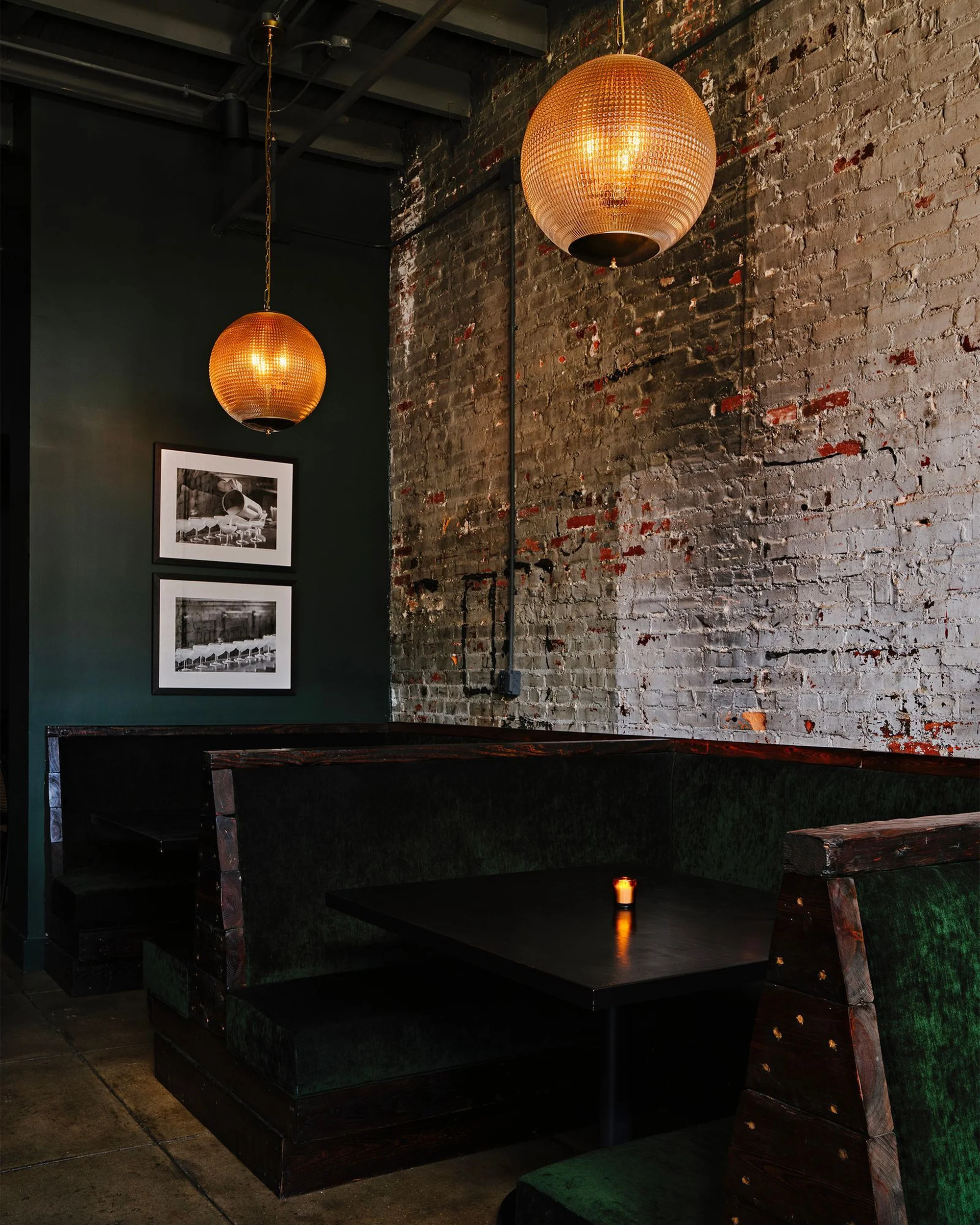 Interior of a dimly lit restaurant with dark green booth seating, a black table with a lit candle, and exposed brick wall with pendant lights.