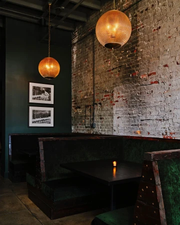 Interior of a dimly lit restaurant with dark green booth seating, a black table with a lit candle, and exposed brick wall with pendant lights.