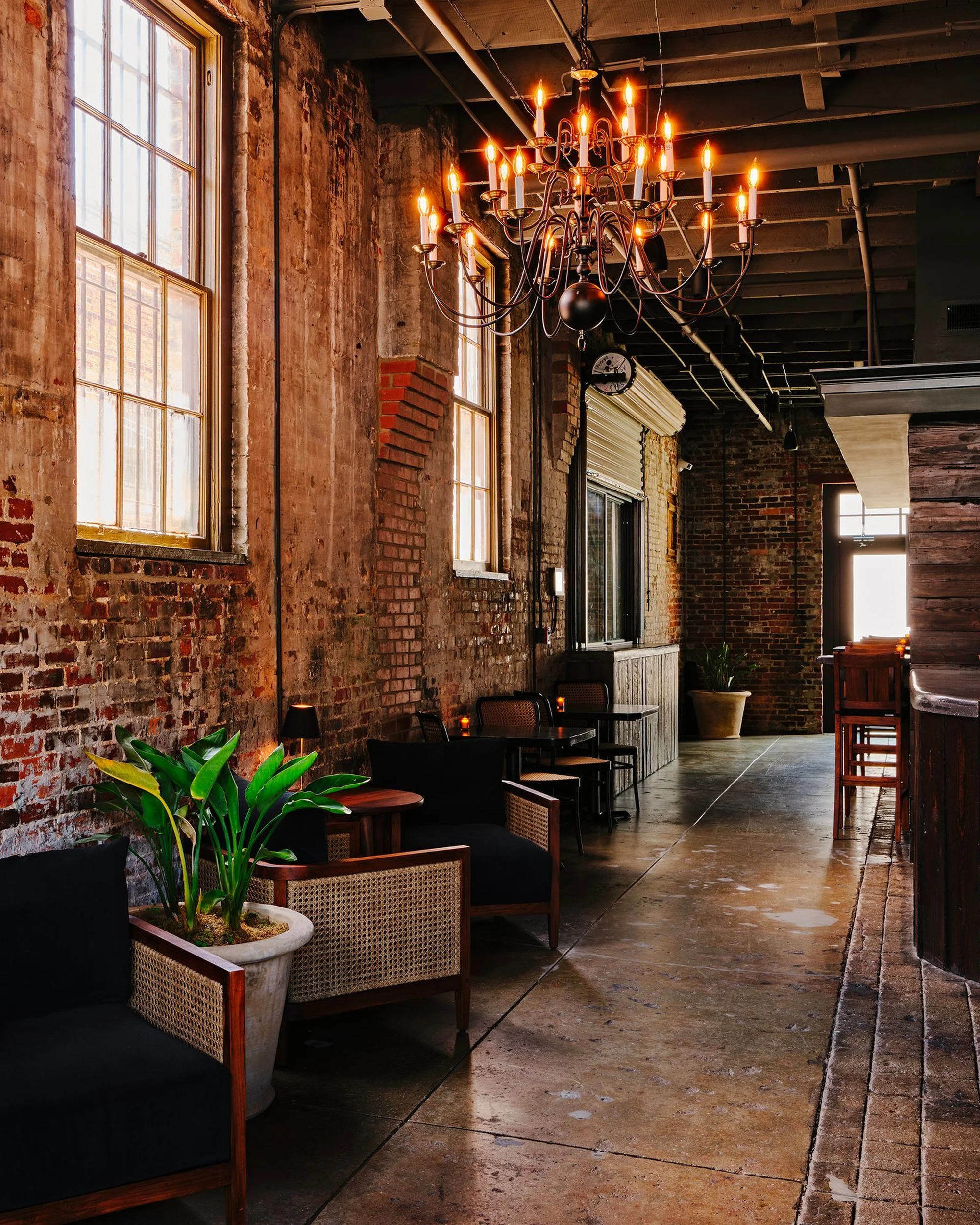 Indoor lounge area featuring chairs, tables, plants, and a chandelier against a brick wall, creating a cozy and dimly lit atmosphere.