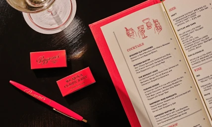 Top-down view of a bright pink restaurant menu, branded pen, matchboxes, and a cocktail glass on a dark tabletop.
