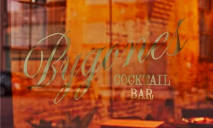 Golden typographic lettering spelling Bygones Cocktail Bar on a glass window reflecting a warm brick building exterior