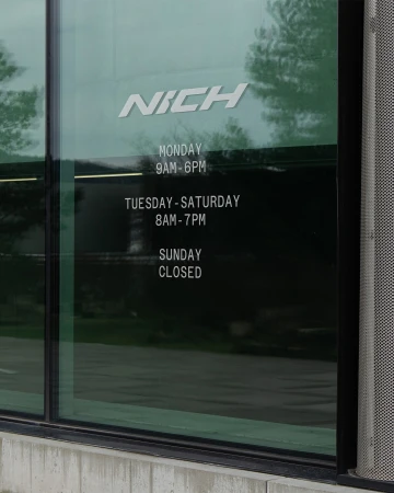 Daytime shot of a building's window displaying business hours for Monday through Sunday, with the business name 'Nich' at the top.