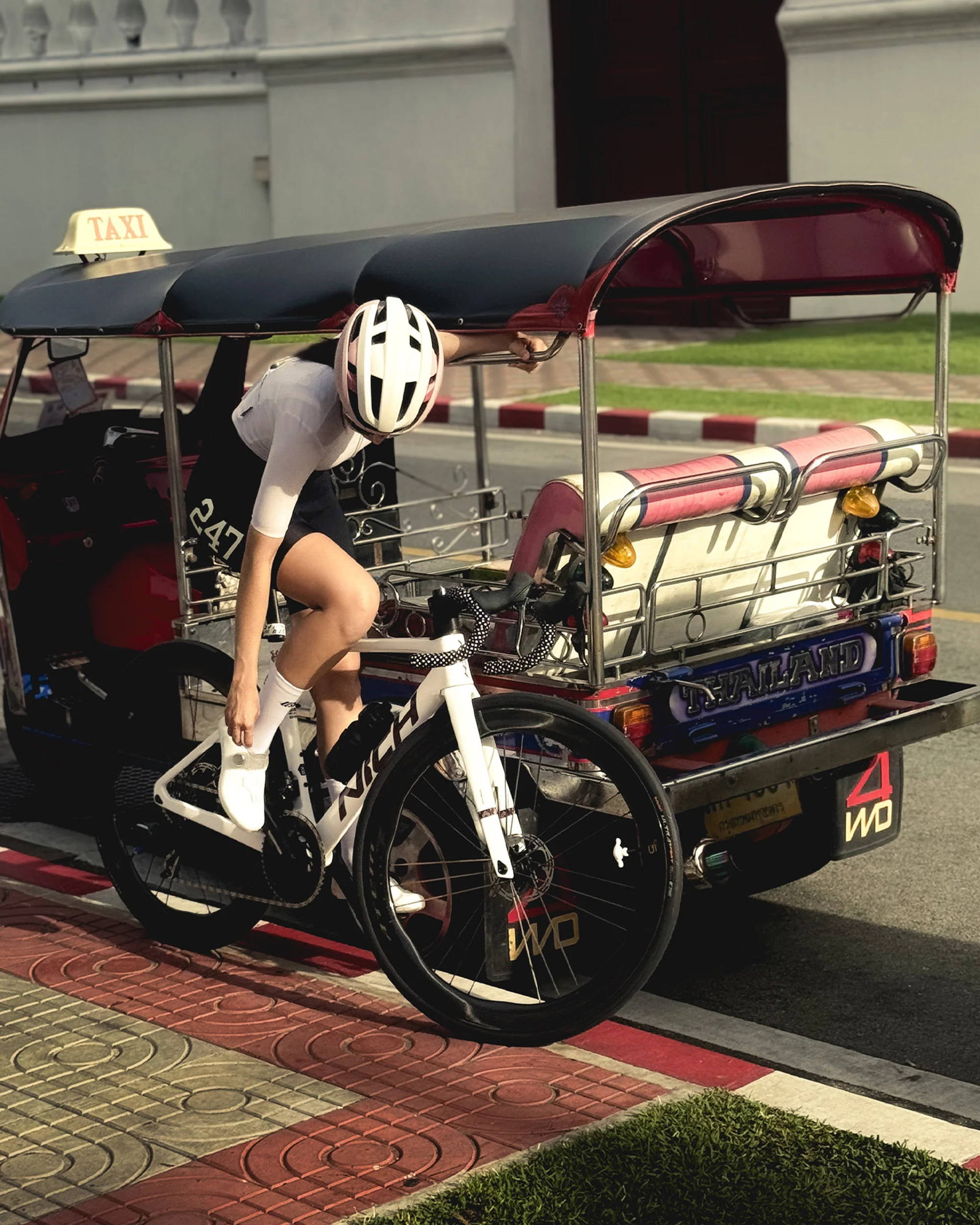 An adult woman in cycling attire is on a bicycle next to a tuk-tuk taxi in an urban setting with neutral light and red, white, and tan colors.