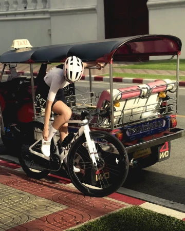 An adult woman in cycling attire is on a bicycle next to a tuk-tuk taxi in an urban setting with neutral light and red, white, and tan colors.