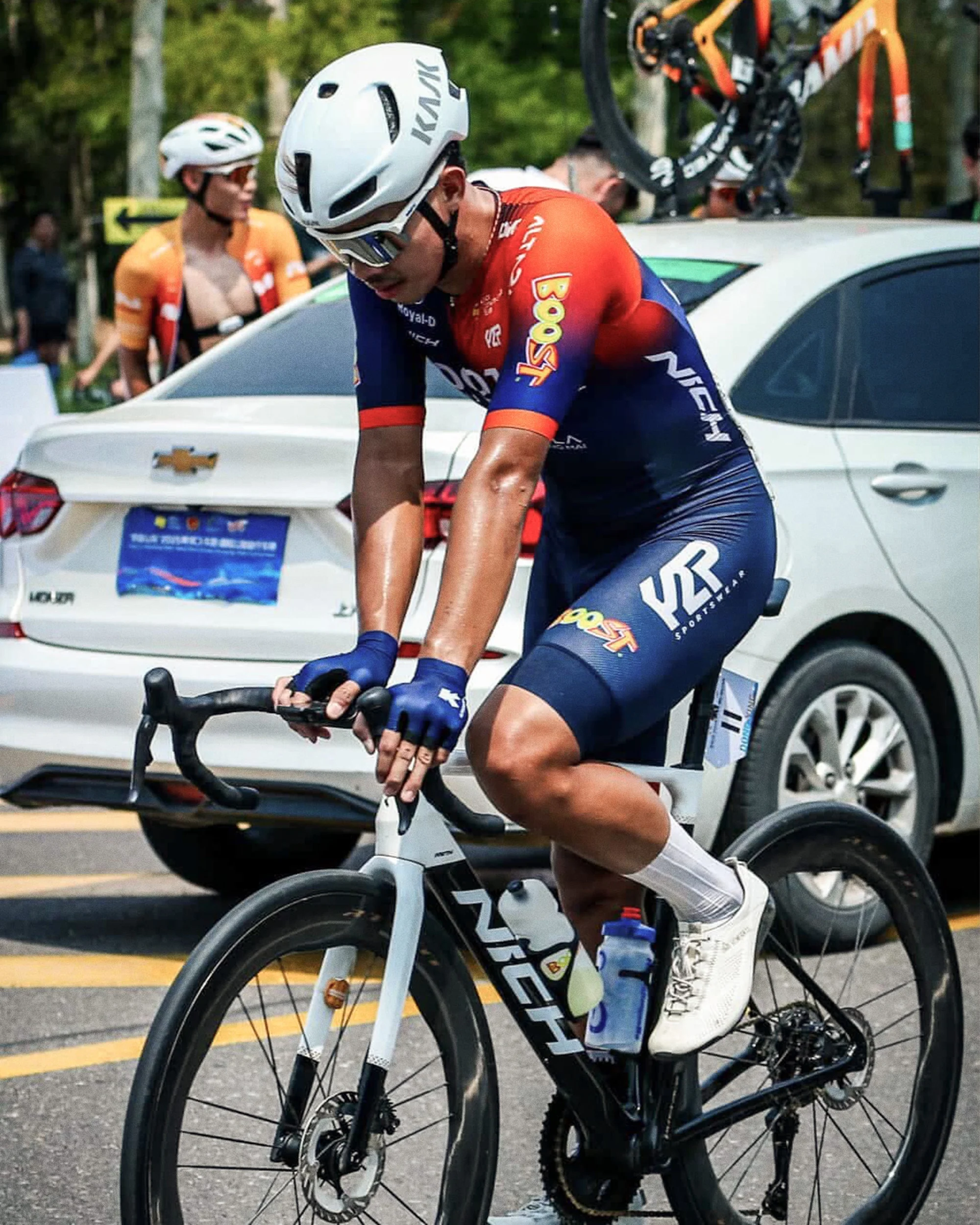 Cyclist in racing gear with helmet on a road bike. Outdoor shot with a car in the background and another racer nearby.