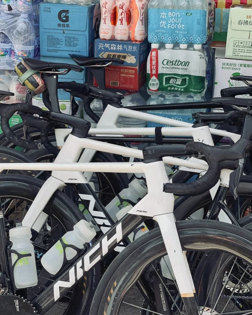 Several road bikes are stacked together outside, with water bottles and beverage containers in the background. Black and white frames, tires visible.