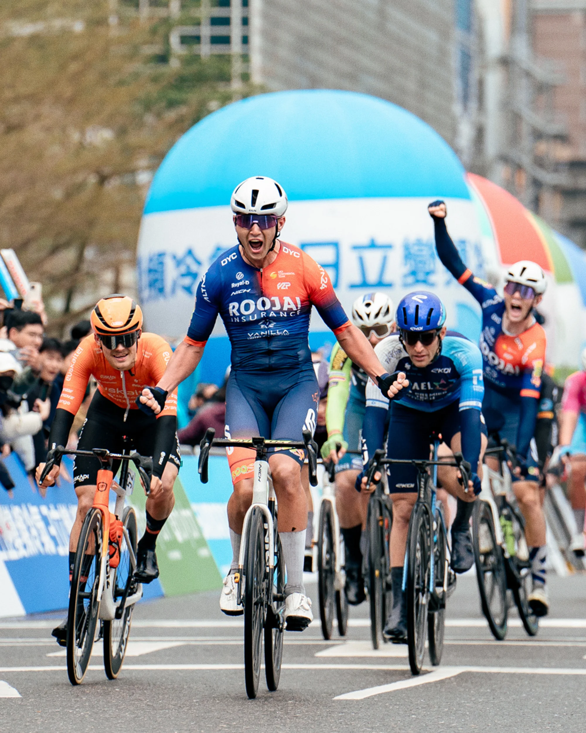 Competitive cyclists racing towards the finish line on a city street, wearing branded sportswear and helmets, cheered on by spectators.