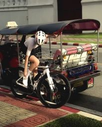 Cyclist in white helmet and jersey leaning against a tuk tuk holding a road bicycle next to a red curb and sidewalk