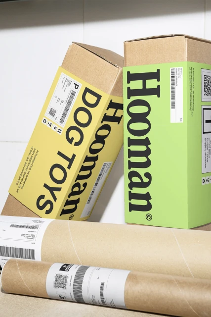 Dog toys subscription boxes from Hooman in yellow and green, arranged on a white shelf with cardboard tubes, in a studio setting.