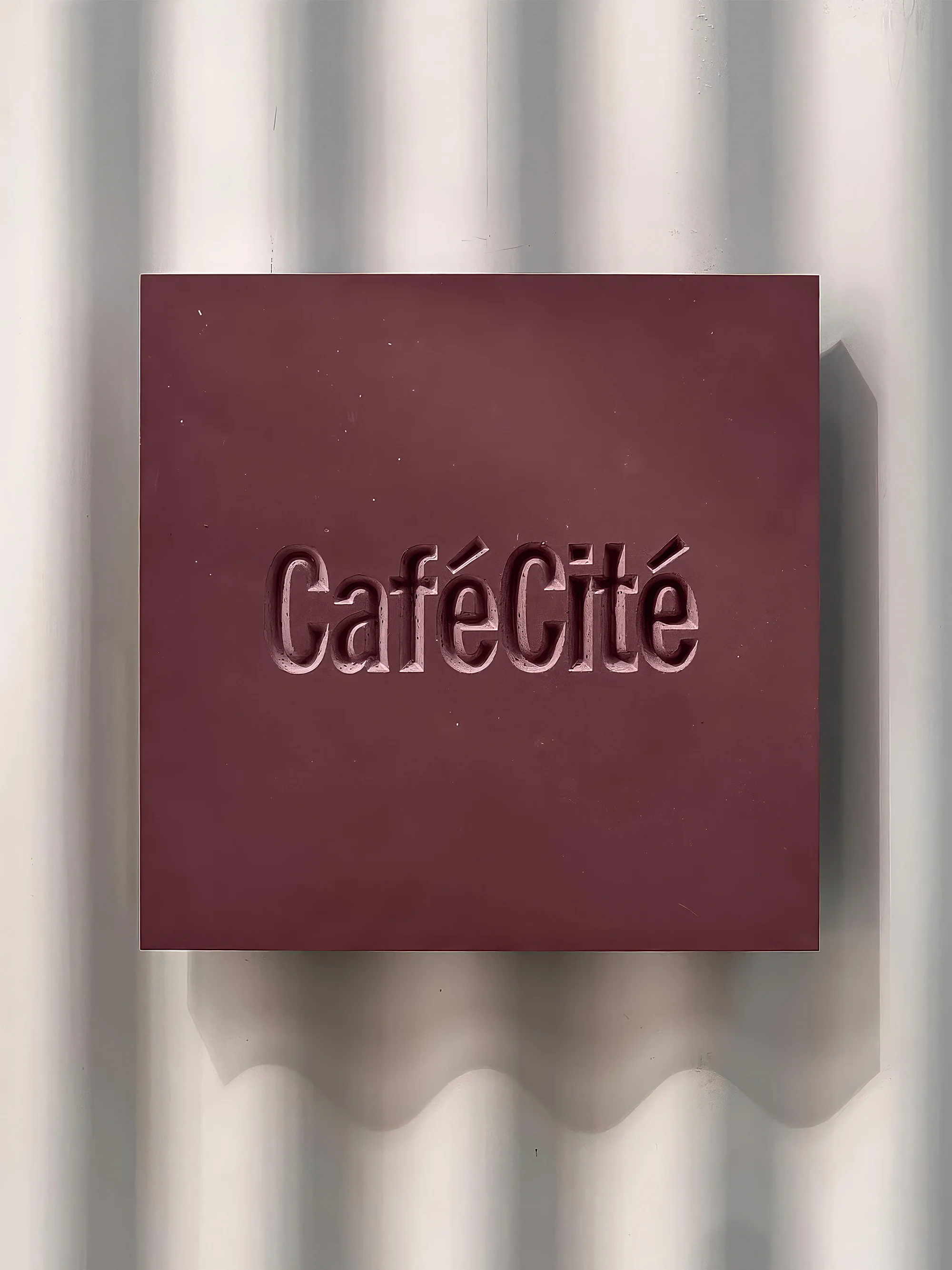 A burgundy sign with the text 'Café Cité' embossed on it is mounted on a white corrugated wall with wavy shadows.