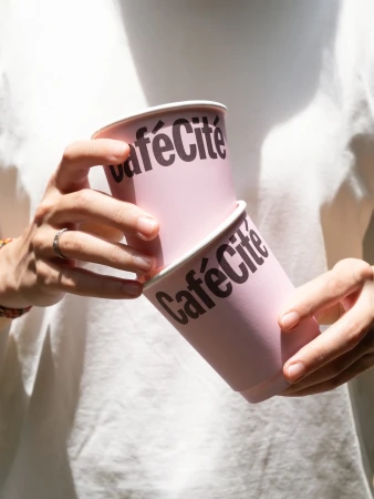 Person holding two pink paper coffee cups with the brand name 'Café Cité' printed on them, against a blurred background.