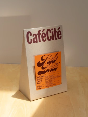 Studio shot of a Café Cité coffee bag on a wooden surface, featuring a custom sticker with the name 'Halo Beriti' on an orange background.