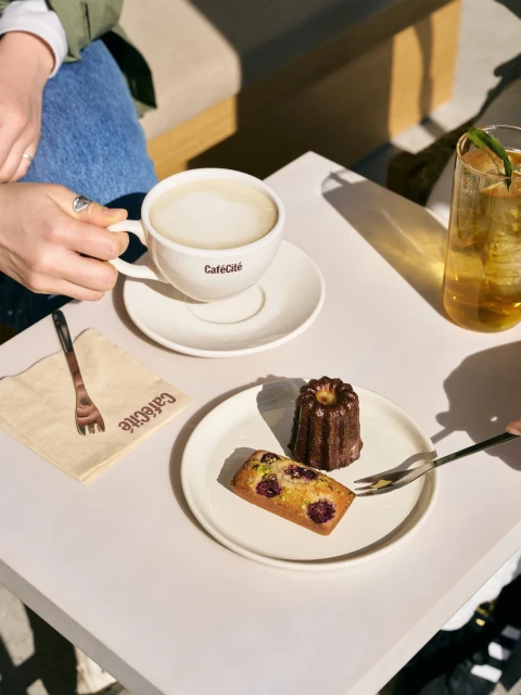 Overhead shot of a table with a latte, pastry, and drink, with two individuals enjoying refreshments in a bright, inviting cafe setting.