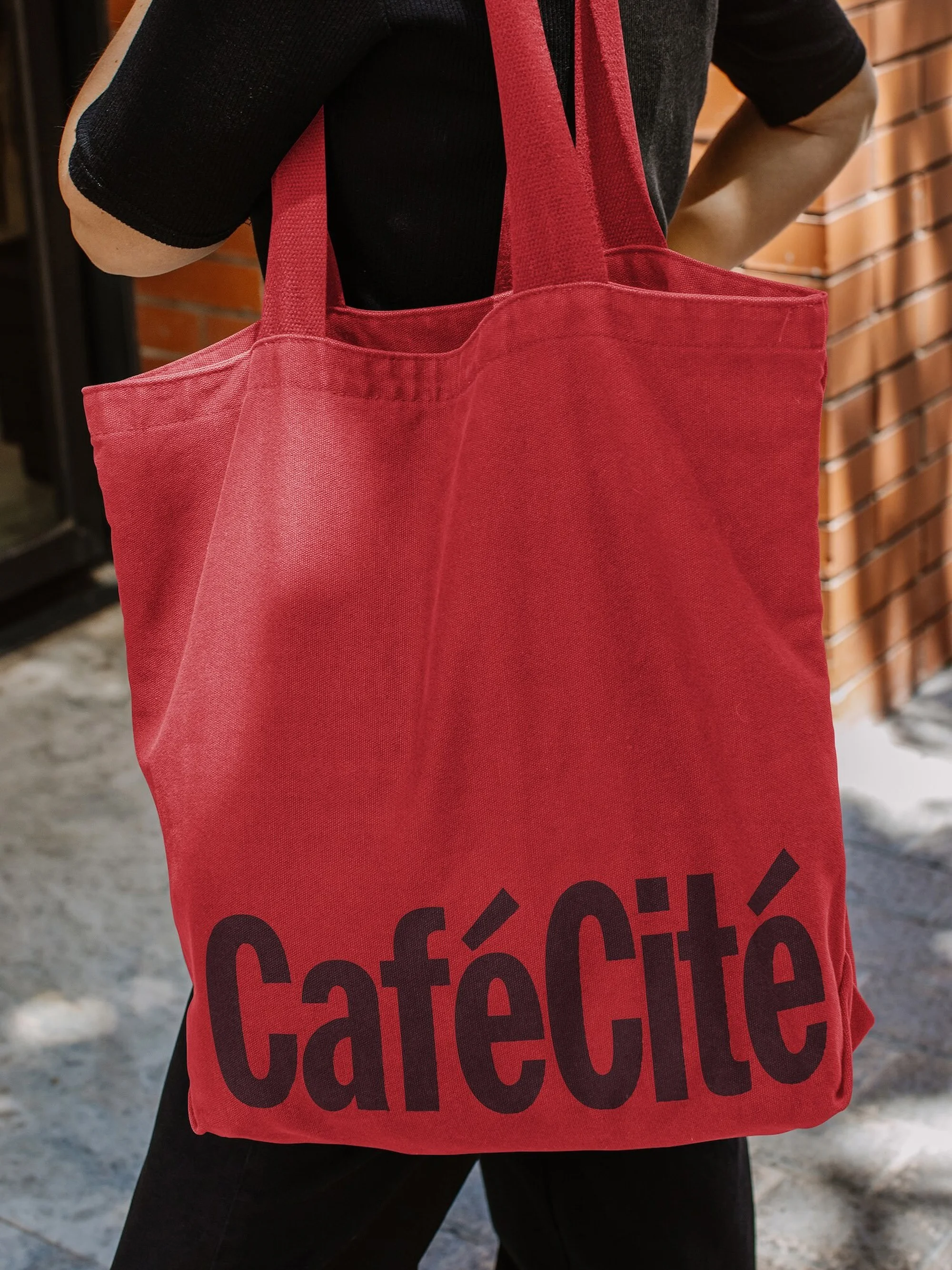 A red canvas tote bag with 'Café Cité' printed on it is slung over a person's shoulder in front of a textured wall outside.