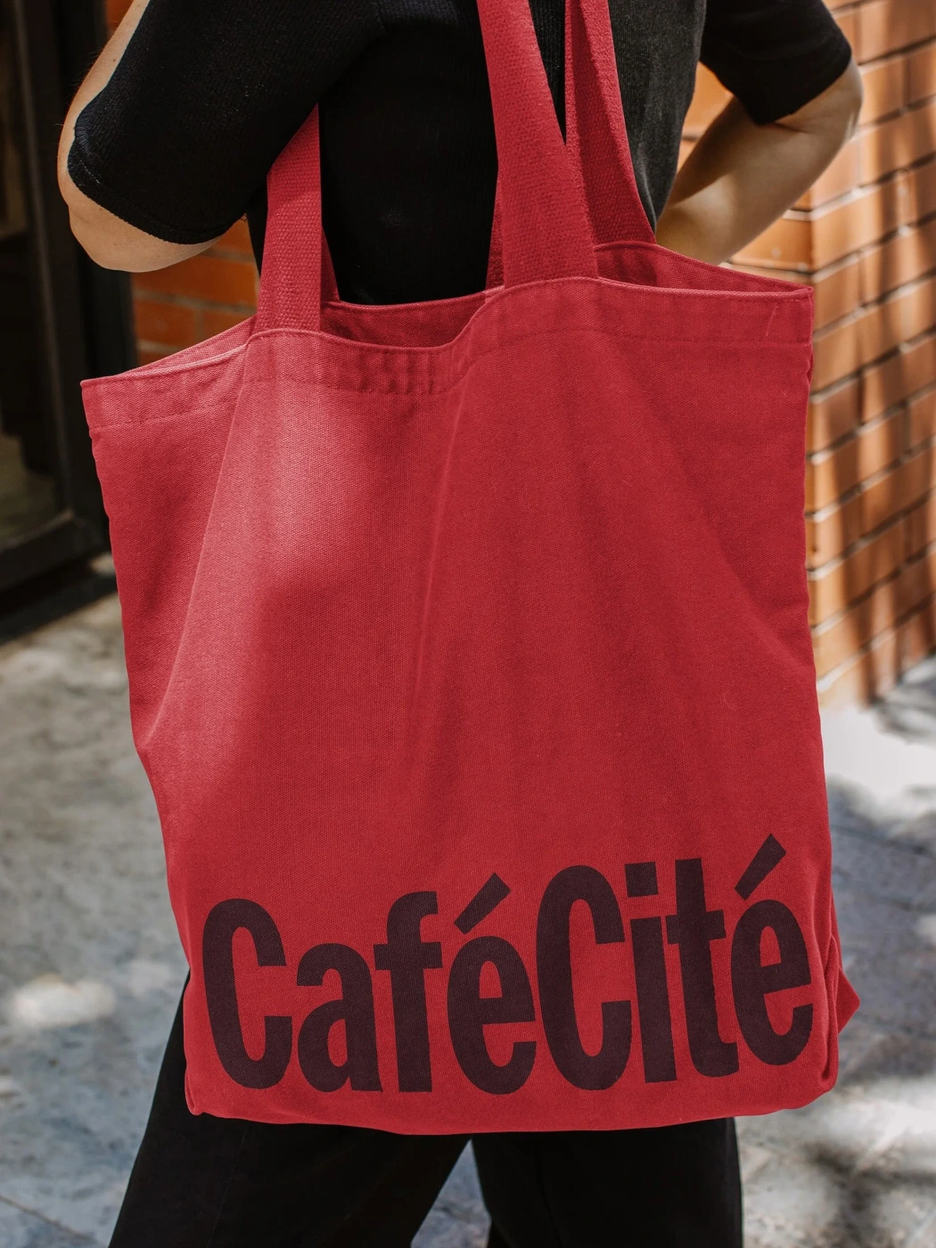 Person carrying a vibrant red canvas tote bag with bold dark typography for CaféCité against an outdoor brick wall background.