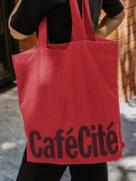Person carrying a vibrant red canvas tote bag with bold dark typography for CaféCité against an outdoor brick wall background.
