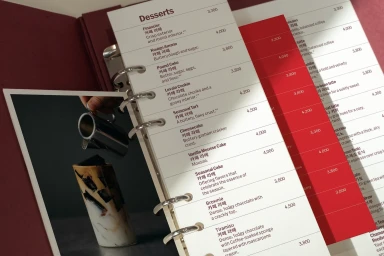 Open ring-bound menu with desserts list in English and Korean, alongside a vibrant red insert and a small inset photo of coffee pouring.