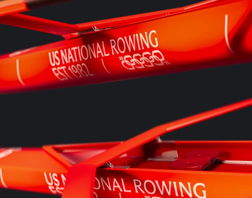 Close-up of an orange rowing scull featuring text and the Olympic rings logo, set against a dark background, focusing on sports equipment.