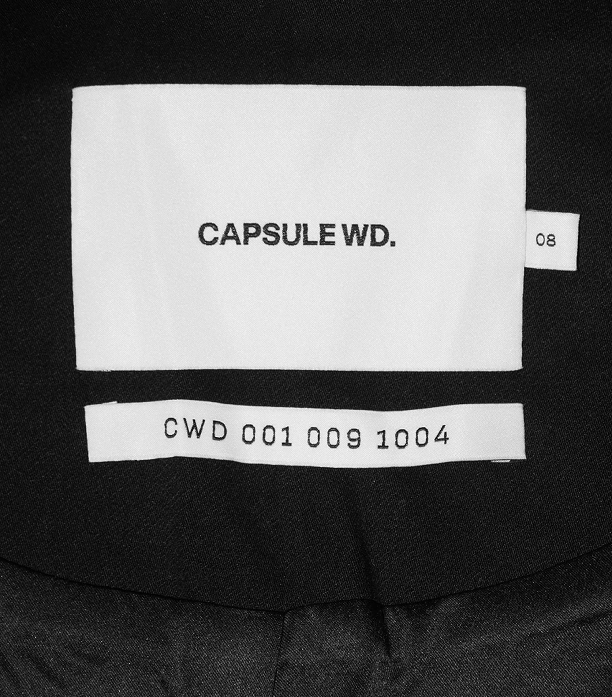 Close-up studio shot of a white garment tag with 'CAPSULE WD.' and 'CWD 001 009 1004' printed in black on a dark garment.