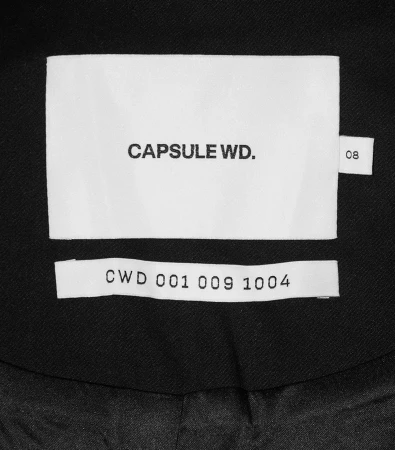 Close-up studio shot of a white garment tag with 'CAPSULE WD.' and 'CWD 001 009 1004' printed in black on a dark garment.