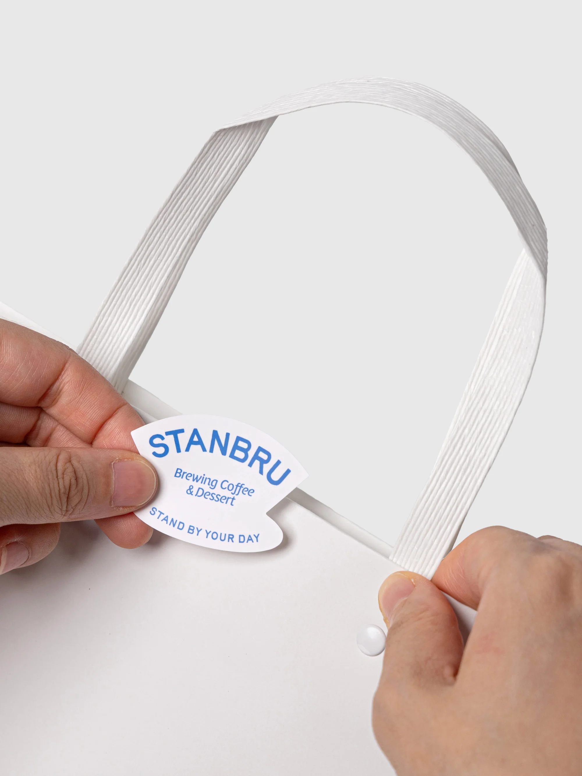 Close-up of a white paper shopping bag held by two hands, showing a blue logo for STANBRU Brewing Coffee & Dessert.