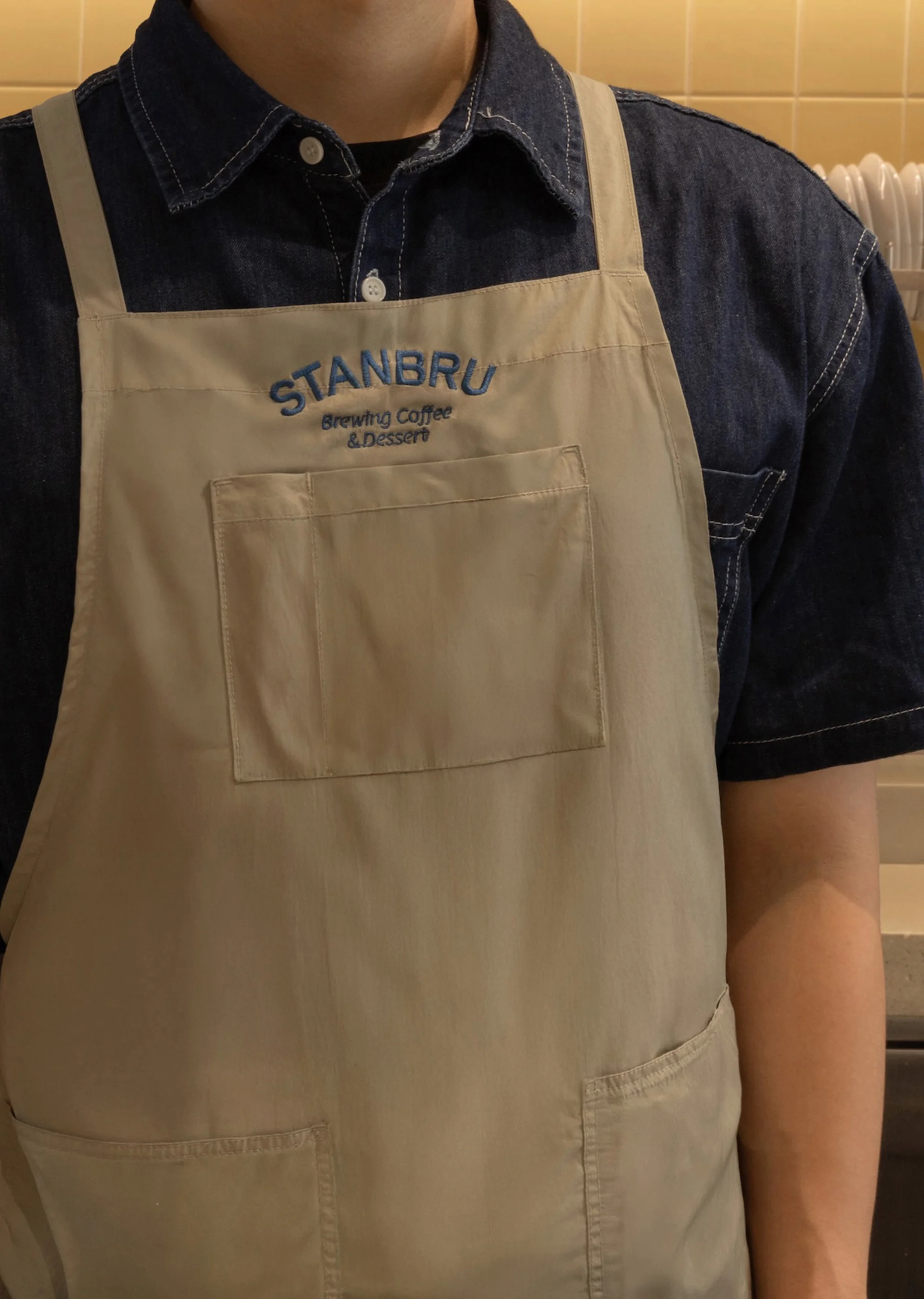 Person wearing a beige apron with 'STANBRU Brewing Coffee & Dessert' embroidered, captured in a medium shot with warm lighting.