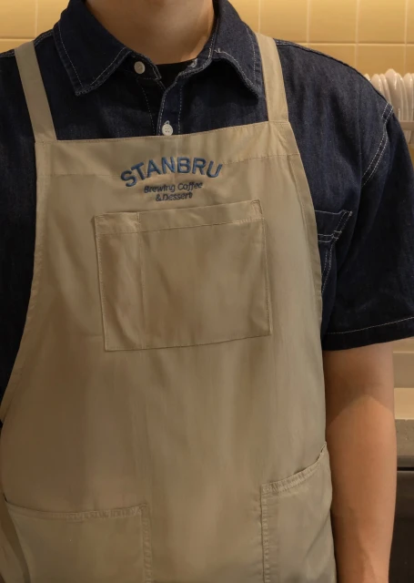 Person wearing a beige apron with 'STANBRU Brewing Coffee & Dessert' embroidered, captured in a medium shot with warm lighting.