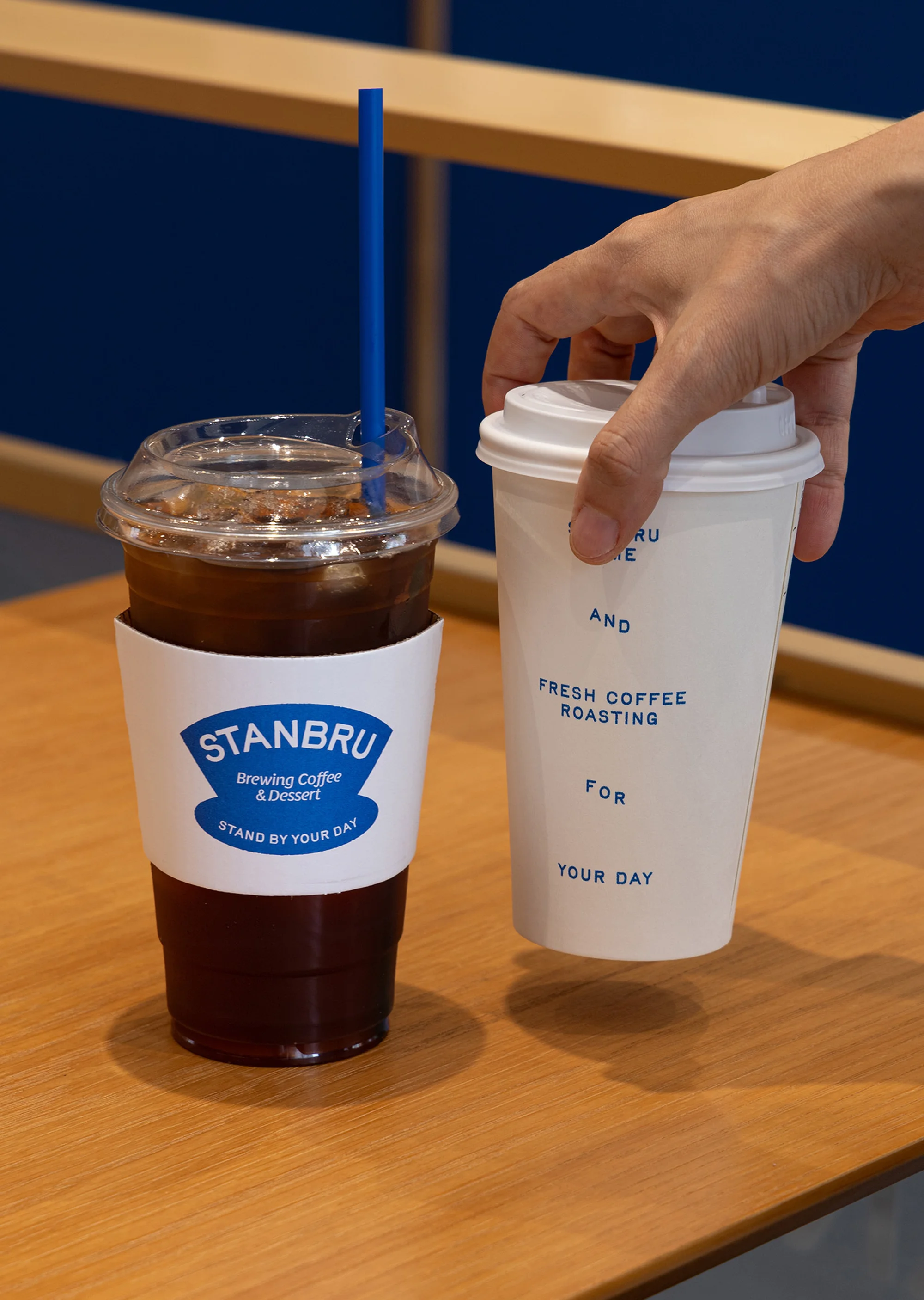 Two Stanbru coffee cups, one iced with a blue straw and one hot, sit on a wooden table against a blue wall, held by a person's hand.