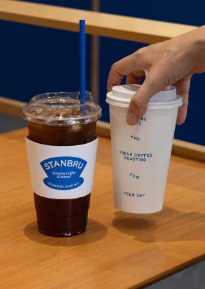 Hand holding a white paper coffee cup next to a plastic iced coffee cup with a blue straw on a wooden table.