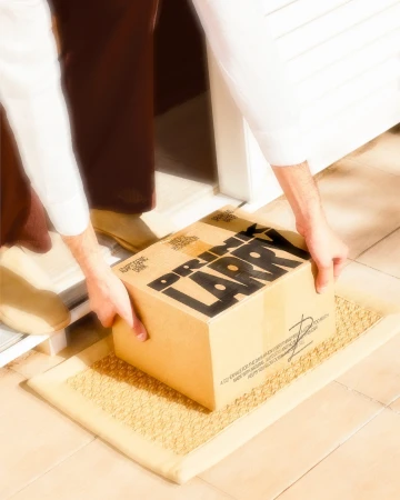 A person is delivering a cardboard box labeled 'Drink Larry' to a house. Tan tiled floor and door. The scene is brightly lit.