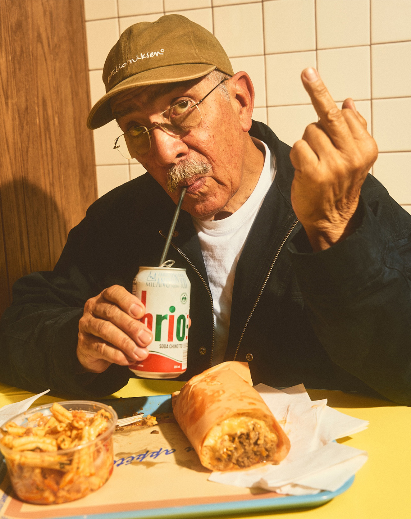 Elderly man in a cap drinks from a can with a straw, flips the bird, with a sandwich and fries on a yellow table.