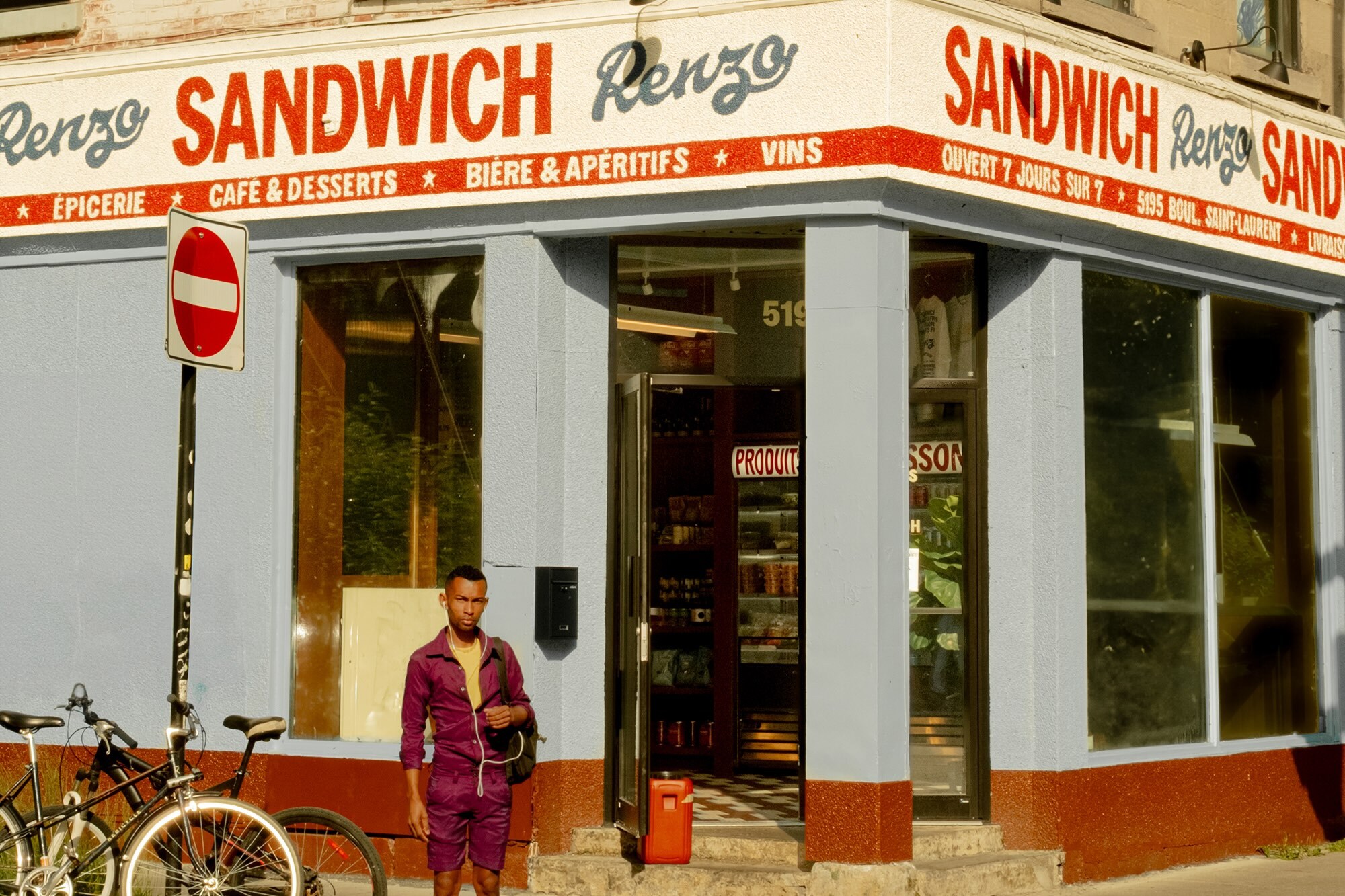 Man in purple jumpsuit stands outside Renzo's sandwich shop next to bicycle on a sunny day, featuring a vintage storefront.