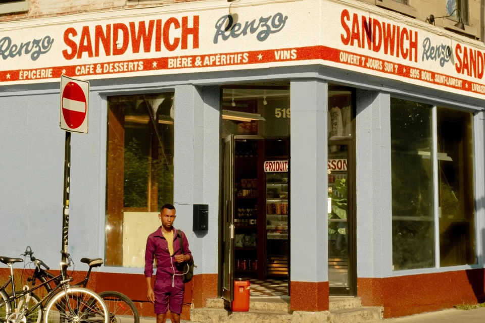 Man in purple jumpsuit stands outside Renzo's sandwich shop next to bicycle on a sunny day, featuring a vintage storefront.