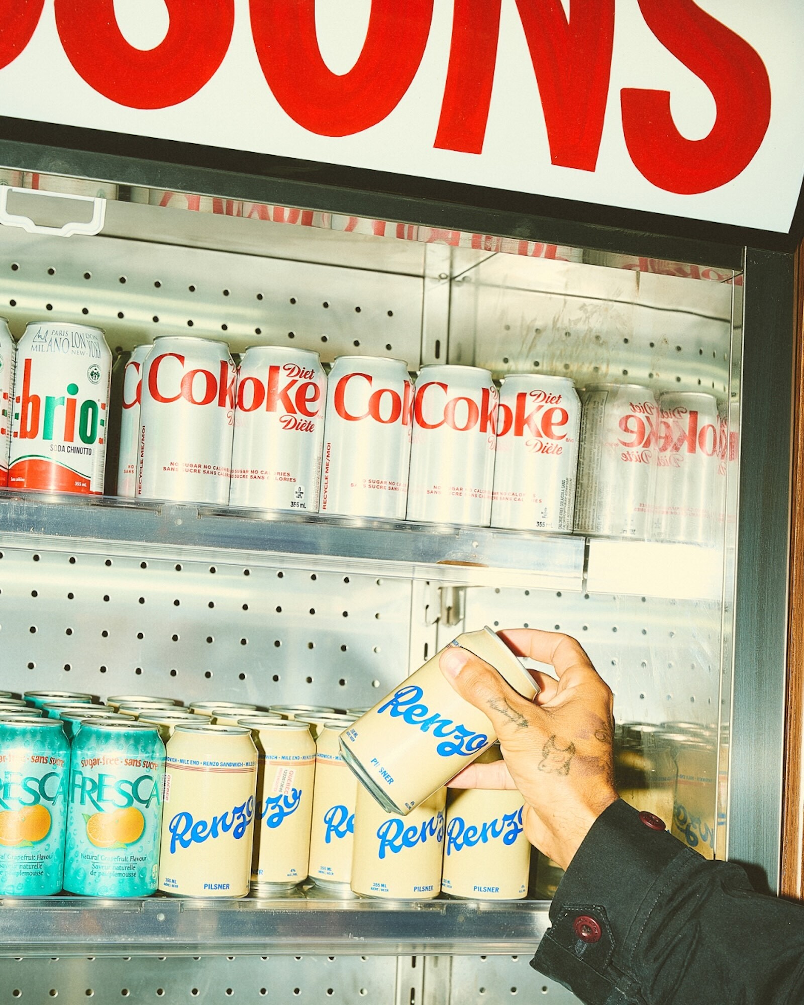 A hand reaches for a can of Renzo soda in a cooler with various soda brands and red text signage at the top. Drinks in background.