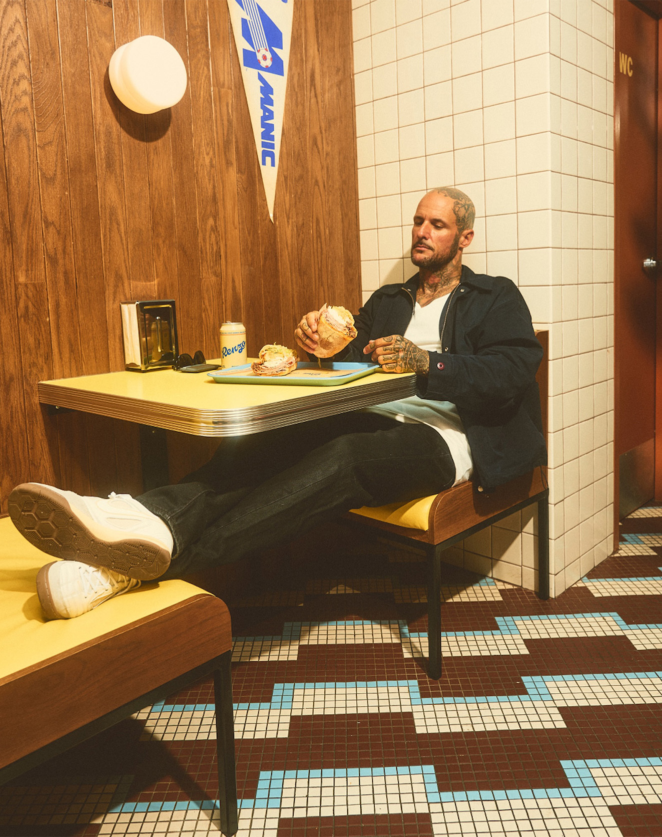 Man with tattoos sits indoors at retro-style table eating sandwich with feet up; walls paneled and tiled; Pennant banner in background.