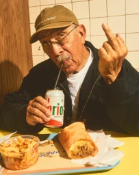Elderly man in a cap drinking from a can and gesturing with his middle finger while seated at a table with food.